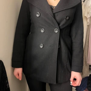 Svart kappa med knappar - Snygg svart kappa med dubbelknäppning och långa ärmar. Klassisk design med knappar både fram och bak för en stilren look. Perfekt för kyligare dagar.