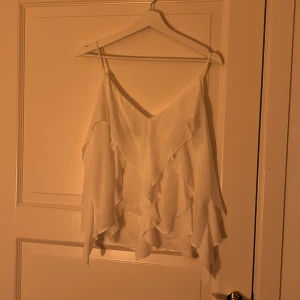 Vit volangtopp från Zara - Säljer en elegant vit topp från Zara med volangdetaljer. Toppen är ärmlös och har tunna axelband, perfekt för en stilren look. Tillverkad i ett lätt och luftigt material.