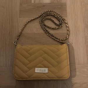 Gul quiltad axelväska från Bebe - Snygg gul axelväska från Bebe med quiltad design och kedjerem. Väskan har en praktisk dragkedja inuti och stängs med en tryckknapp. Perfekt för att ge en färgklick till din outfit!