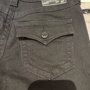 Svarta bootcut jeans från True Religion - Snygga svarta bootcut jeans från True Religion med midwaist passform i storlek 27. De har dekorativa fickor med knappar och en klassisk design. Perfekta för en stilren look. Haft på mig 1 gång men dom var tyvärr stora på mig. Pris mellan 1000-1100 pågrund av att dom aldrig använts och är som helt nya