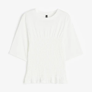 Vit topp med smock - Säljer en stilren vit topp med trekvartsärm. Toppen har en smockad detalj framtill som ger en fin struktur och en lätt volang vid nederkanten. Perfekt för en enkel och elegant look💗Skriv för frågor!🤗💕