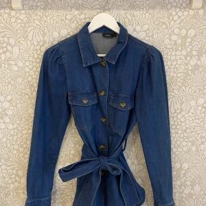 Blå jeansjacka med knytband - Snygg blå jeansskjorta från Kappahl med knappar och knytband i midjan. Jackan har långa ärmar och två bröstfickor med knappar. 