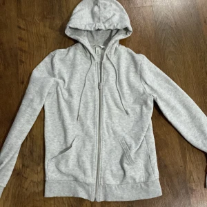 Grå hoodie från H&M - Säljer en stilren grå hoodie från H&M. Den har en dragkedja framtill och en praktisk huva med dragsko. Perfekt för en avslappnad look. Passar bra till både jeans och mjukisbyxor.