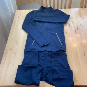 Mörkblå träningsjacka och byxor från Nike - Säljer en mörkblå träningsjacka och matchande byxor från Nike. Jackan har dragkedja och två fickor med vita detaljer. Byxorna har en elastisk midja med dragsko och en vit Nike-logga på benet. Perfekt för träning eller en avslappnad dag! Riktigt skönt sett! 