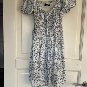 Blommig klänning från BikBok - Charmig vit klänning med blått blommönster från BikBok. Den har korta ärmar och en söt knytning framtill. Smockdetaljer ger en bekväm passform och klänningen är perfekt för varma dagar. Då den är för liten för mig så kan jag tyvärr inte visa den på men annars är den så mycket finare på