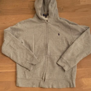 Grå hoodie från Ralph Lauren - Säljer en klassisk grå hoodie från Ralph Lauren med dragkedja och broderad logga på bröstet. Tröjan har långa ärmar och en bekväm huva. Storleken är en 14-16 år så ungefär 160 i barn storlek 