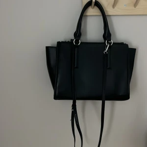 Svart axelväska med dragkedja  - Snygg svart axelväska i syntetmaterial med praktisk dragkedja upptill. Väskan har stilrena detaljer och en justerbar axelrem för bekväm användning. Det går även att ta bort banden om man vill💗