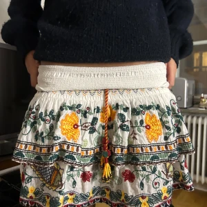 Vit broderad kjol från zara  - Charmig vit kjol med färgglada broderier och volangdetaljer. Kjolen har en elastisk midja och ett vackert blommönster i grönt, gult och rött. Perfekt för en bohemisk look!