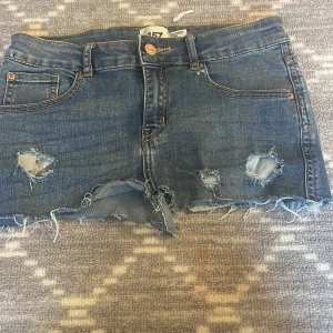 Blå jeansshorts från 157 - Snygga blå jeansshorts från 157 med slitna detaljer och fransig kant. De har en klassisk femficksdesign och knappgylf. Perfekta för en avslappnad stil.