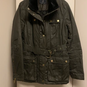 Olivgrön jacka från Stockholm - Snygg olivgrön jacka från Stockholm med svart fuskpäls på kragen. Jackan har både dragkedja och knappar framtill samt ett bälte i midjan för en justerbar passform. Praktiska fickor med knappar och detaljer i guldton ger en stilren look.