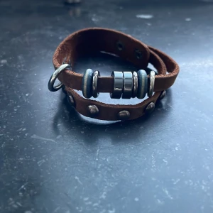 Brunt läderarmband med metallringar - Brunt läderarmband med flera metallringar och nitar. Armbandet har en justerbar knäppning med tryckknappar.
