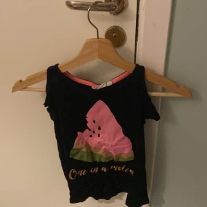 Svart topp med vattenmelontryck från KappAhl - Säljer en svart topp från KappAhl med ett färgglatt vattenmelontryck och texten 'One in a melon'. Toppen har korta ärmar och en rund halsringning. Perfekt för en avslappnad stil.