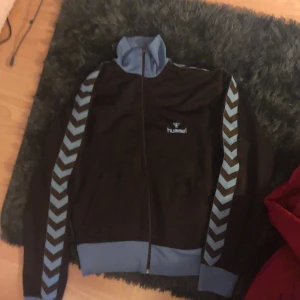 Blå fullzip tröja från Hummel hör av dig för mer - Säljer en blå tröja från Hummel med ljusblå detaljer. Jackan har dragkedja och långa ärmar med det klassiska Hummel-mönstret i ljusblått längs ärmarna. Perfekt för sportiga aktiviteter eller en avslappnad stil.