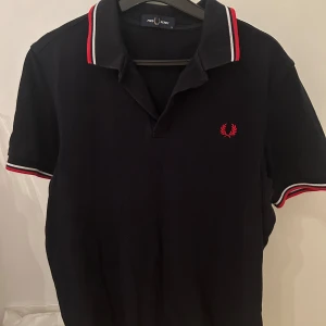 Svart pikétröja från Fred Perry - Säljer en klassisk svart pikétröja från Fred Perry med röda och vita detaljer på krage och ärmslut. Tröjan har en broderad röd logotyp på bröstet och korta ärmar. Perfekt för en stilren look. Storlek m men har krympt i tvätten så passar S