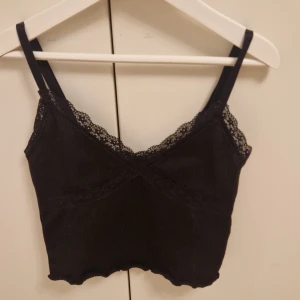 Svart ribbad linnetopp med spetsdetaljer - Snygg svart ribbad linnetopp med spetsdetaljer längs kanterna. Toppen har smala justerbara axelband och en v-ringad design som ger en elegant touch. Perfekt för att styla med olika outfits. 