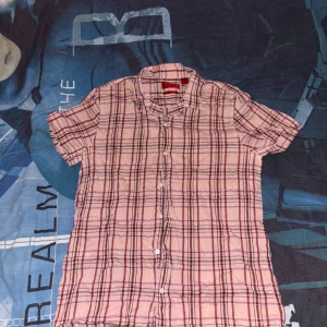 Rosa kortärmad skjorta från Hugo Boss - Säljer en snygg rutig skjorta från Hugo Boss i rosa och bruna nyanser. Skjortan är kortärmad och har knappar framtill. Perfekt för en avslappnad stil. Jätteskön till sommaren och väldigt trendig. Välbevarad och inte så använd.