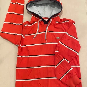 Röd randig hoodie från Lacoste - Säljer en snygg röd hoodie från Lacoste med vita och svarta ränder. 