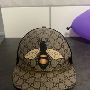 Beige keps med bi-motiv från Gucci - Snygg beige keps från Gucci med ett coolt bi-motiv på framsidan. Kepsen har ett klassiskt GG-mönster och justerbar passform. Perfekt för att ge din outfit en lyxig touch. Skick 8 av 10
