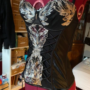 svart korsett topp med tre storleksalternativ - svart korsettliknande topp med gulddetaljer och tre nivåer av hyskor baktill. har strumpebandshållare samt axelband som är lätta att ta bort. genomskinlig / mesh i ryggen. kupa 70C. obs små förslitningar på öglan som axelbanden sitter fast på