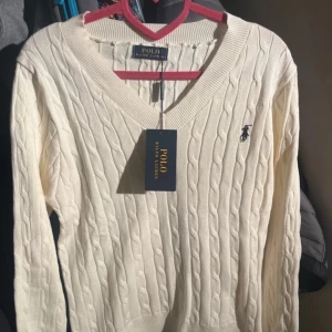 Vit kabelstickad tröja från Polo Ralph Lauren  - Säljer en stilren vit kabelstickad tröja från Polo Ralph Lauren. Tröjan har en v-ringad hals och det klassiska Polo-logotypen broderad på bröstet. 
