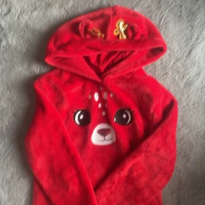 Röd hoodie med djurmotiv - Mysig röd hoodie med ett sött djuransikte på framsidan och dekorativa detaljer i guld på huvan. Perfekt för att hålla sig varm och bekväm. Den har långa ärmar och är gjord i ett mjukt material.