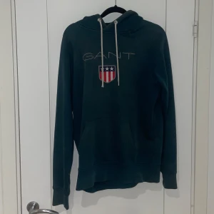 Mörkgrön hoodie från GANT - Snygg mörkgrön hoodie från GANT med logotyp och emblem på bröstet. Tröjan har en klassisk känguruficka och justerbar huva med snören. Perfekt för en avslappnad stil.