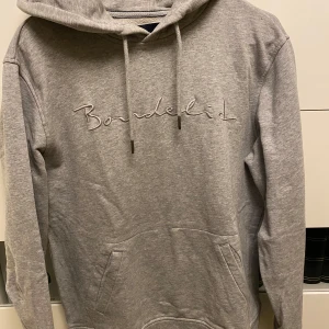 Grå hoodie från Bondelid - Säljer en stilren grå hoodie från Bondelid med broderad logga på bröstet. Tröjan har en klassisk känguruficka och  huva med snören. Perfekt för en avslappnad look.