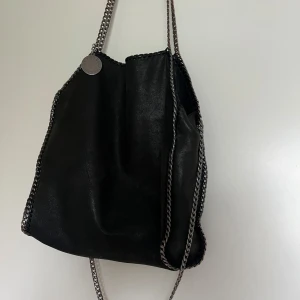 Liknande Stella McCartney väska  - Snygg svart axelväska som liknar Stella McCartney. Den har kedjedetaljer och en lila insida. Jag vet inte huruvida den är äkta eller inte då den är köpt begagnad. Hör gärna av dig innan du trycker på Köp nu! 💛