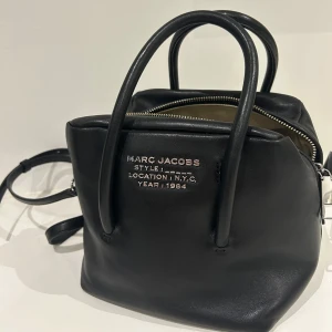Svart handväska från Marc Jacobs - Snygg svart handväska från Marc Jacobs med textdetaljer på framsidan. Väskan har två handtag och en avtagbar axelrem för flexibilitet. Det finns även en till väska innuti som går att ta ut. Medföljer även en dustbag till väskan