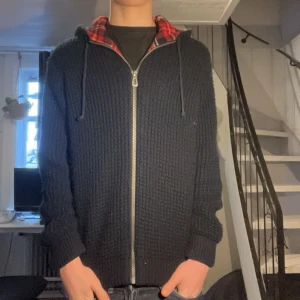 Stickad cardigan - Säljer en mörkblå cardigan med rutig insida i rött och vitt. Den har en dragkedja framtill och en mysig huva. Perfekt för kyliga dagar!
