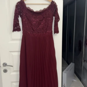 Vinröd spetsklänning - Elegant vinröd långklänning med spetsdetaljer och trekvartsärmar. Klänningen har en vacker spetsöverdel och en plisserad kjol som ger en fin siluett. Perfekt för festliga tillfällen.