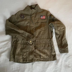 Olivgrön jacka från Esprit - Säljer en olivgrön jacka från Esprit med patches på bröstet och ärmarna. Jackan har en hög krage och praktiska fickor framtill. 