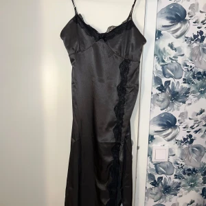 Svart långklänning med spetsdetaljer - Elegant svart långklänning med smala axelband och vacker spets längs kanterna. Klänningen har en figurnära passform och är tillverkad i ett glansigt material som ger en lyxig känsla. Perfekt för speciella tillfällen. Klänningen har en lätt slit sträcker sig ända till knäna. Mörkgrå med lätta glitter i material 