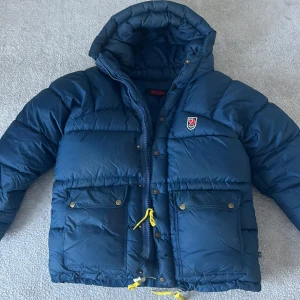 Blå dunjacka från Fjällräven - Säljer en blå dunjacka från Fjällräven med huva och dragkedja. Jackan har knappar och två stora fickor framtill med gul snörning. Perfekt för kalla dagar.