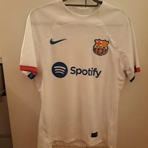 Nike Barcalona tröja - Säljer en vit fotbollströja från Nike med FCB-emblem och Spotify-logga på framsidan. Tröjan har korta ärmar med röd och blå detalj vid ärmsluten. Baksidan har UNHCR-logga. Perfekt för fotbollsfans!