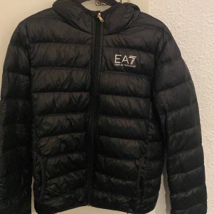 Svart jacka från Emporio Armani - Snygg svart dunjacka från Emporio Armani EA7 med huva och dragkedja framtill. Perfekt för kyliga dagar med sin stilrena design och bekväma passform.