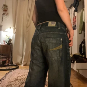 Mörkblå jeans med broderad detalj - Säljer ett par mörkblå jeans med en cool broderad detalj på bakfickan. De har en loose passform och är lågmidjade, perfekta för en avslappnad stil. Märket är Evisu, känt för sina unika designinslag. W 33, L 32. Funkar för mig som bär storlek 38-40 i byxor.