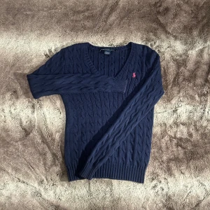 Ralph Lauren - Säljer en stilren mörkblå kabelstickad tröja från Ralph Lauren. Tröjan har en v-ringad design och ett broderat rosa logotypmärke på bröstet. Perfekt för en klassisk och avslappnad look.