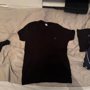 Svart t-shirt från Moncler - Snygg svart t-shirt från Moncler med rund hals och korta ärmar. Den har en diskret logga på bröstet som ger en stilren look. Perfekt för en casual outfit. Lite kortare vid ärmarna som gör att du ser mer biff ut.