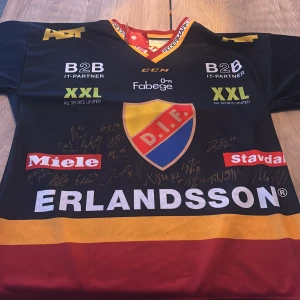 Mörkblå  hockeytröja från CCM med DIF-logga signerad - Säljer en mörkblå hockeytröja från CCM med DIF-logga och flera sponsormärken. Tröjan har en gul och röd krage och är täckt med autografer. Perfekt för hockeyfans!
