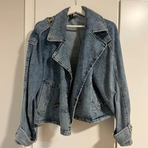 Blå jeansjacka med knappar - Snygg blå jeansjacka med dubbelknäppning och bred krage. Jackan har en avslappnad passform och långa ärmar med uppvikta muddar. Perfekt för en cool och casual look.