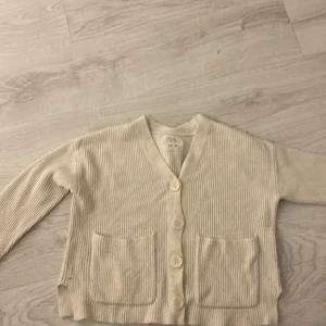 Beige ribbad kofta från Zara - Säljer en mysig beige kofta från Zara i storlek 140. Koftan har en ribbad struktur med knappar framtill och två praktiska fickor. Perfekt för kyliga dagar!
