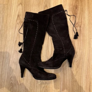 Bruna höga mockastövlar med klack - Eleganta bruna stövlar i mocka med hög klack och dekorativa snören med tofsar. Varsamt använda, inga defekter förutom lite på snörena, men de syns knappt