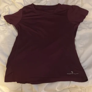 Vinröd t-shirt från Ronhill - Snygg vinröd t-shirt från Ronhill, perfekt för träning. Den har korta ärmar och är tillverkad i ett lätt och andningsbart material. Diskret logga på framsidan.