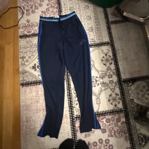 Blå träningsbyxor från Adidas - Snygga blå träningsbyxor från Adidas med ljusblå detaljer och logga på benet. Perfekta för träning eller en avslappnad dag. Byxorna har en bekväm passform och elastisk midja.