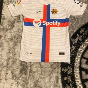 Vit Barcelona fotbollströja med Ansu Fati 10 - Snygg vit Barcelona fotbollströja från Nike med Ansu Fati och nummer 10 på ryggen. Tröjan har korta ärmar och ett korsmönster i rött och blått på framsidan. Perfekt för fans av laget och spelaren! Storlek 176/S