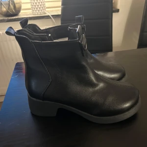 Svarta boots med dragkedja - Snygga svarta boots i syntetmaterial med dragkedja på sidan och dekorativa spännen. Perfekta för höst och vinter med en bekväm passform och stilren design.