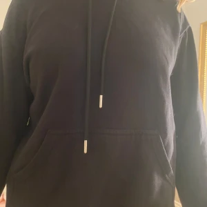 Svart hoodie med ficka - Säljer en klassisk svart hoodie med känguruficka och justerbar dragsko i huvan. Perfekt för en avslappnad stil.