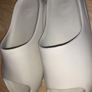 Yeezy slides - Säljer ett par bekväma beige sandaler från Adidas. De har en stilren design med öppen tå och en räfflad sula för extra grepp. Perfekta för avslappnade dagar.   Storlek 44 om man har 43 kan man växa in i de. Ifall man inte vill betala frakt så kan jag möta upp er i Stockholm 