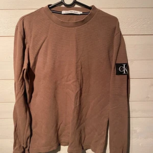 Beige tröja från Calvin Klein Jeans - Säljer en stilren beige tröja från Calvin Klein Jeans med långärmad design och ribbad struktur. Tröjan har en diskret CK-logga på ärmen och rund halsringning. Perfekt för en avslappnad look.